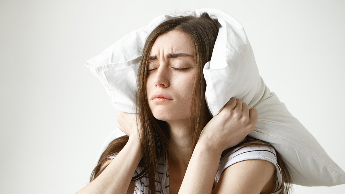 Pretty student girl in striped t-shirt trying to have some sleep, sitting in bedroom with pillow on her head, closing eyes, having tired painful look, irritated with annoying sound. Negative emotions